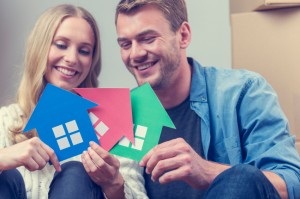 Couple with 3 house symbols – choice concept. They are sitting down, smiling, both casually dressed and holding green, blue and red house symbols which could represent also environmental conservation or home ownership, or property development.